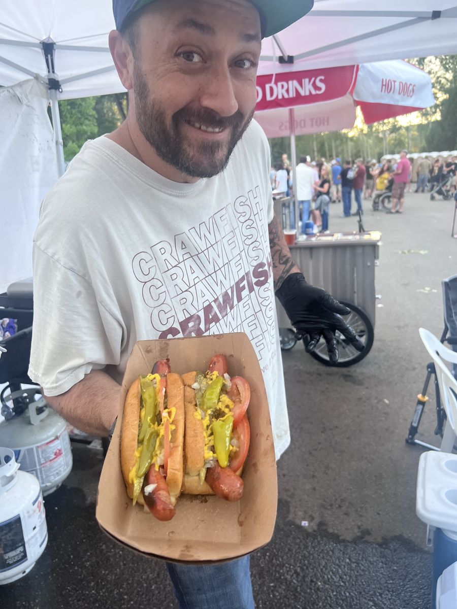 A smiling House of Hogs volunteer holding a tray of fully-loaded Chicago dogs, with the festival crowd behind him